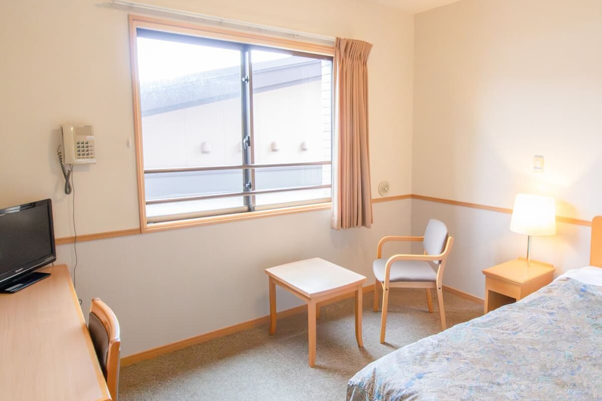 A hotel room with a bed, a desk, a chair, a TV, a window with curtains, and a lamp.
