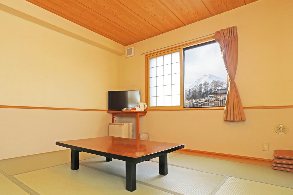 A room with tatami flooring, a low table, a television, and a window with a view of a mountain.