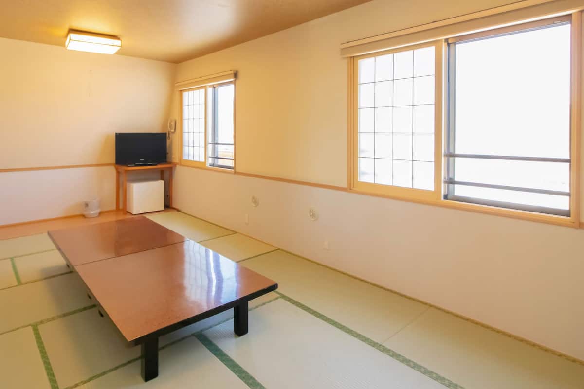 A room with tatami flooring, a low table, a television on a stand, and large windows.