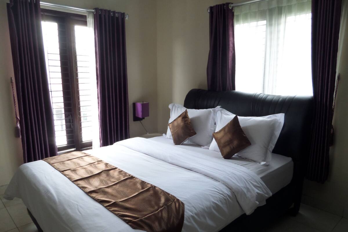 A hotel room with a large bed, dark curtains, a headboard, and a small purple lamp.