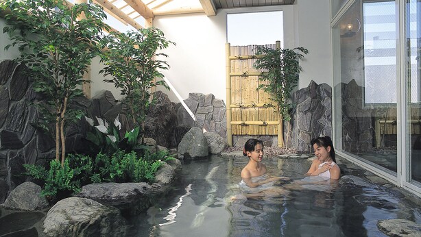 Un área de aguas termales con dos personas en el agua, rodeada de rocas y vegetación, bajo una estructura de techo de madera.