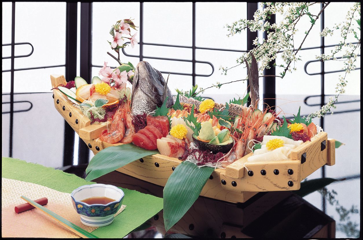 A wooden boat filled with various seafood and flowers, placed on a table with a green mat and a small bowl of soy sauce.