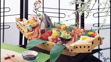 A wooden boat filled with various seafood and flowers, placed on a table with a green mat and a small bowl of soy sauce.