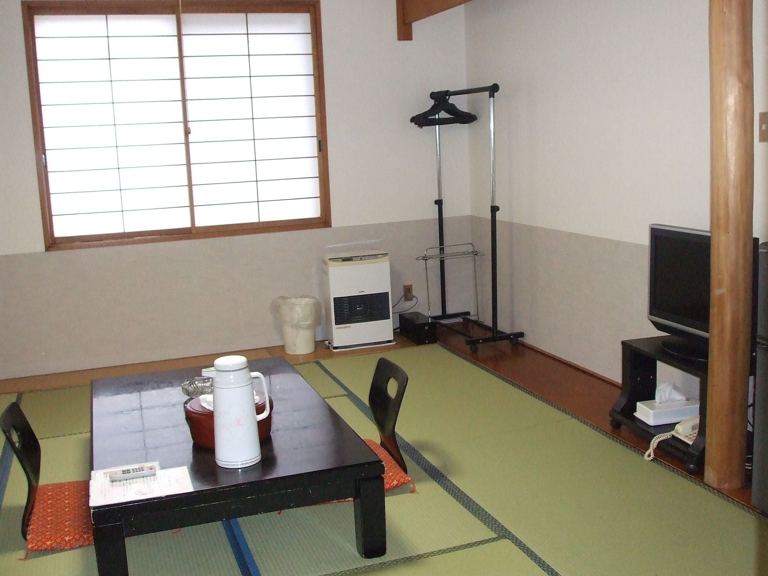 A room with a tatami mat floor, a table with a kettle and a remote control, a heater, a TV, and a hanging clothes rack.