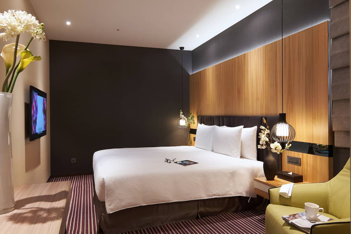 A hotel room with a large bed, a bedside table with a lamp, a small table with a vase of flowers, and a green armchair.