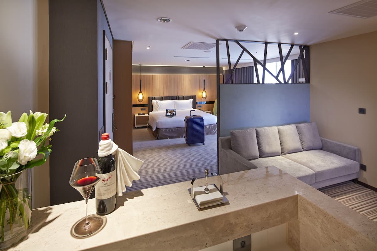 A hotel room with a bed, a sofa, a minibar, a vase with flowers, and a wine glass.