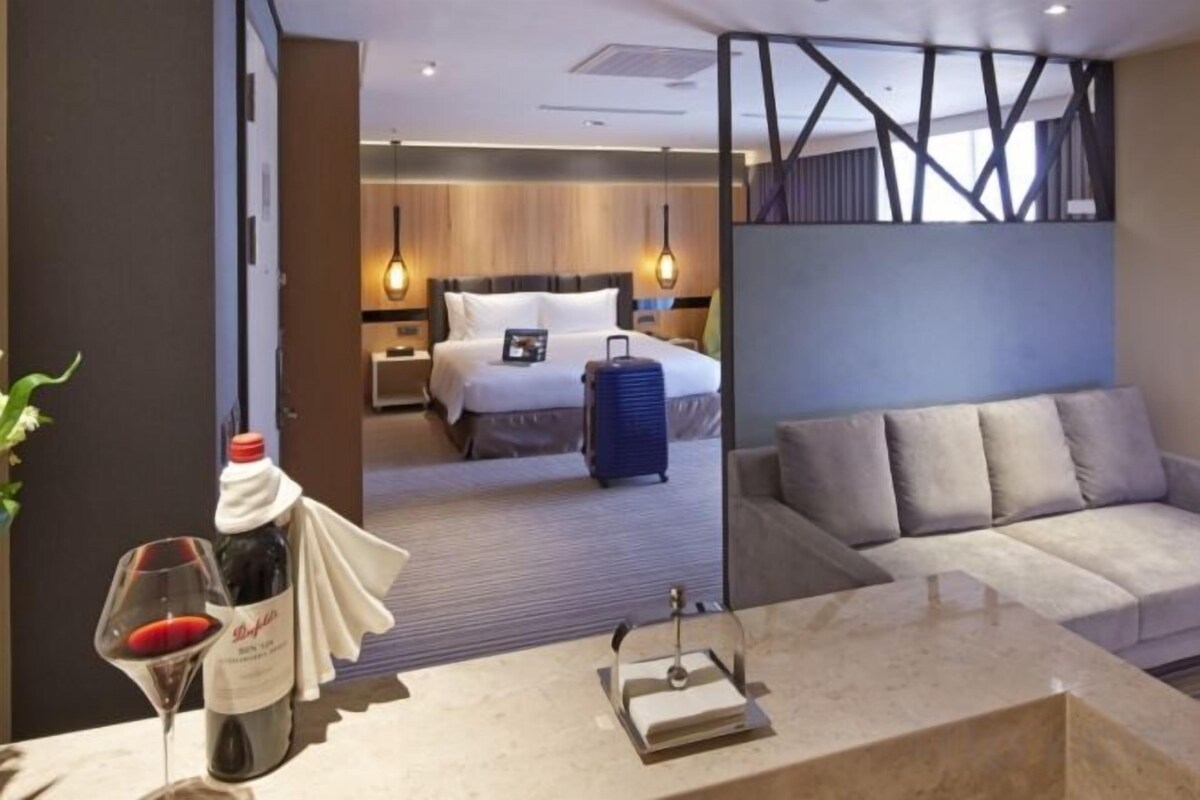 A hotel room with a bed, a sofa, a desk with a bottle and glasses, and a suitcase.