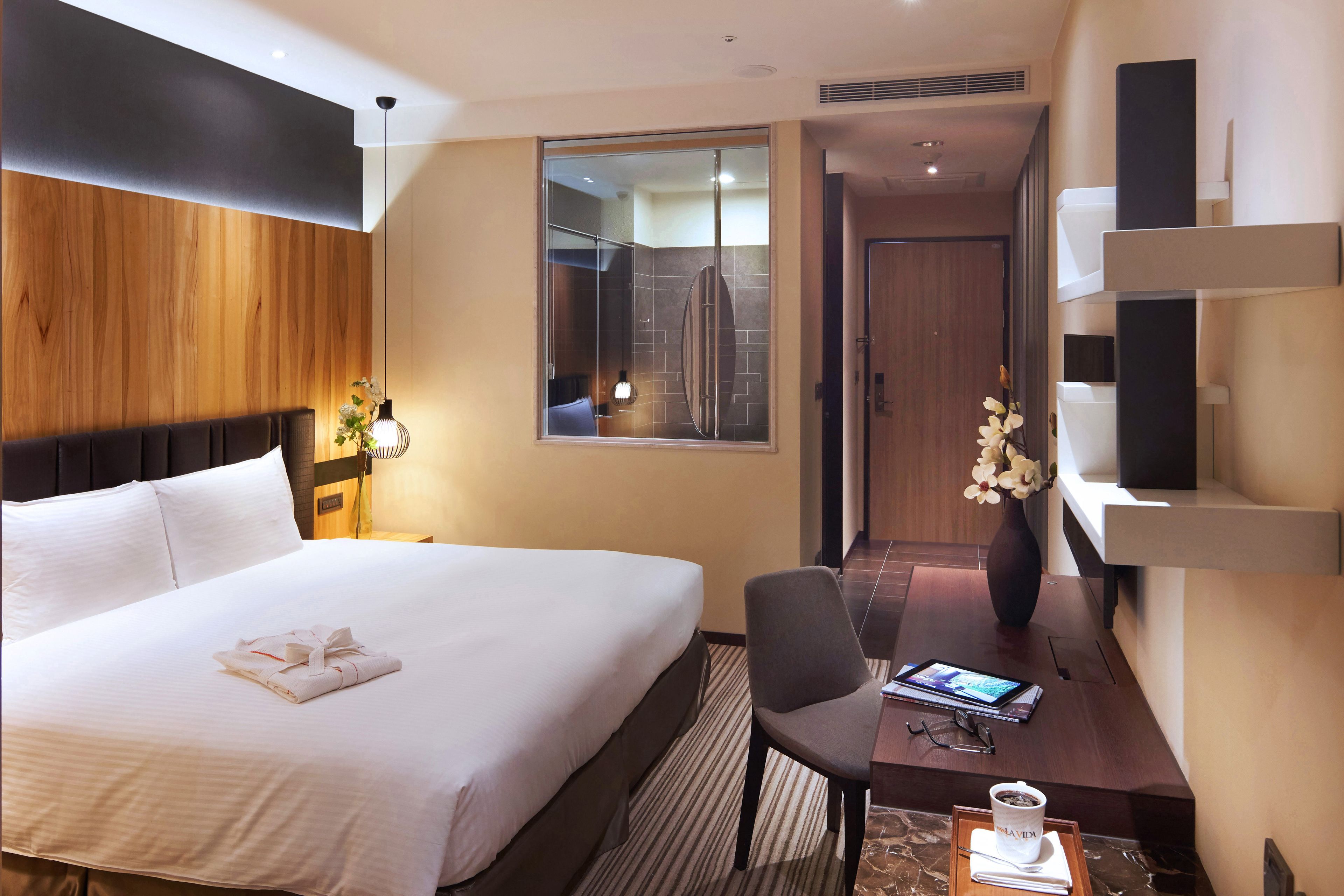 Quarto de hotel com uma cama grande, uma escrivaninha com cadeira, um vaso com flores e vista para o banheiro através de uma porta de vidro.