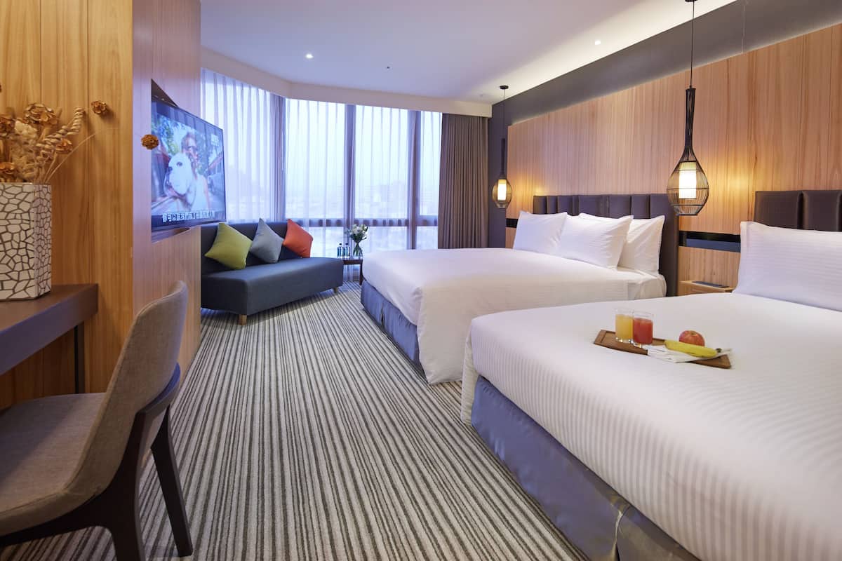 A hotel room with two beds, a TV, a desk with a chair, and a view of the city through large windows.