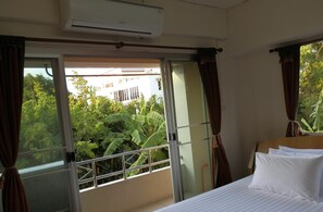 A hotel room with a bed, curtains, an air conditioning unit, and a view of greenery outside.