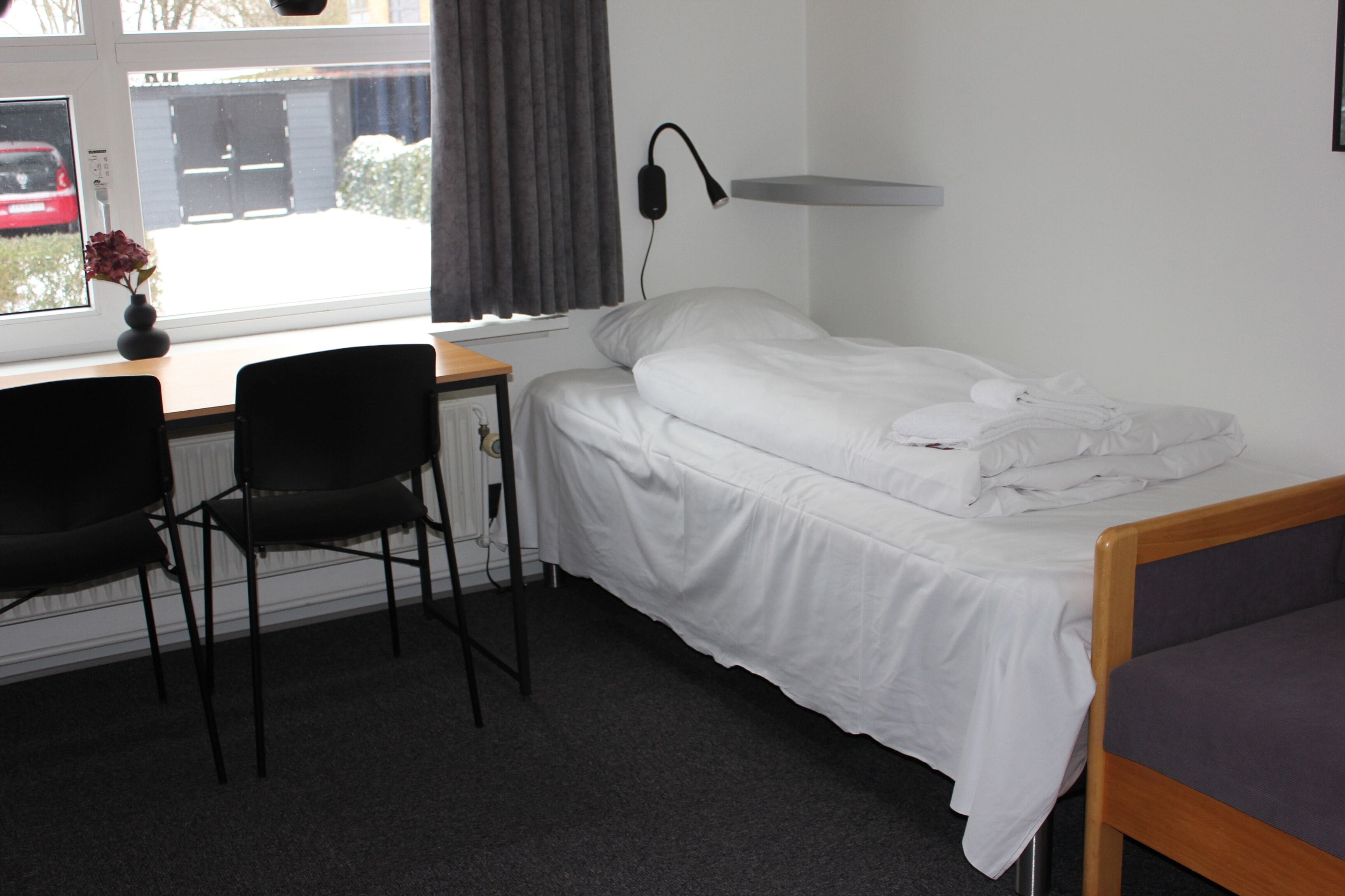 A hotel room with a bed, a desk with a chair, a lamp, and a window with a view of the outside.