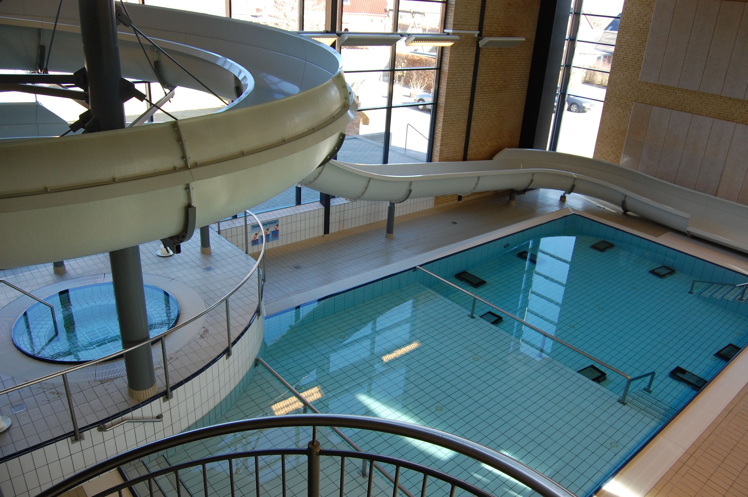 A large indoor swimming pool with a central slide, surrounded by tiled walls and a clear glass barrier.