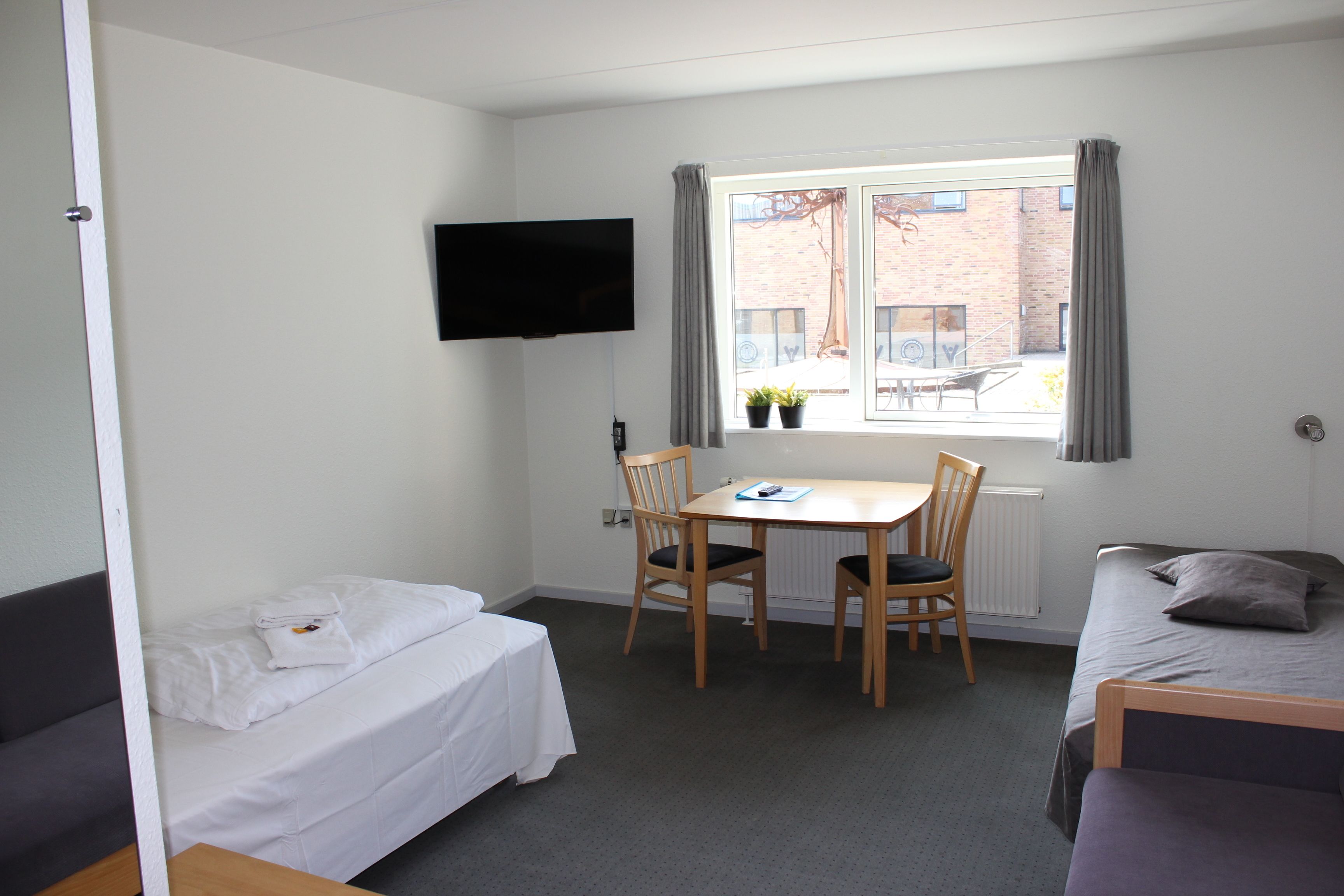 A small, clean room with a bed, a desk with two chairs, a TV on the wall, and a window with curtains.