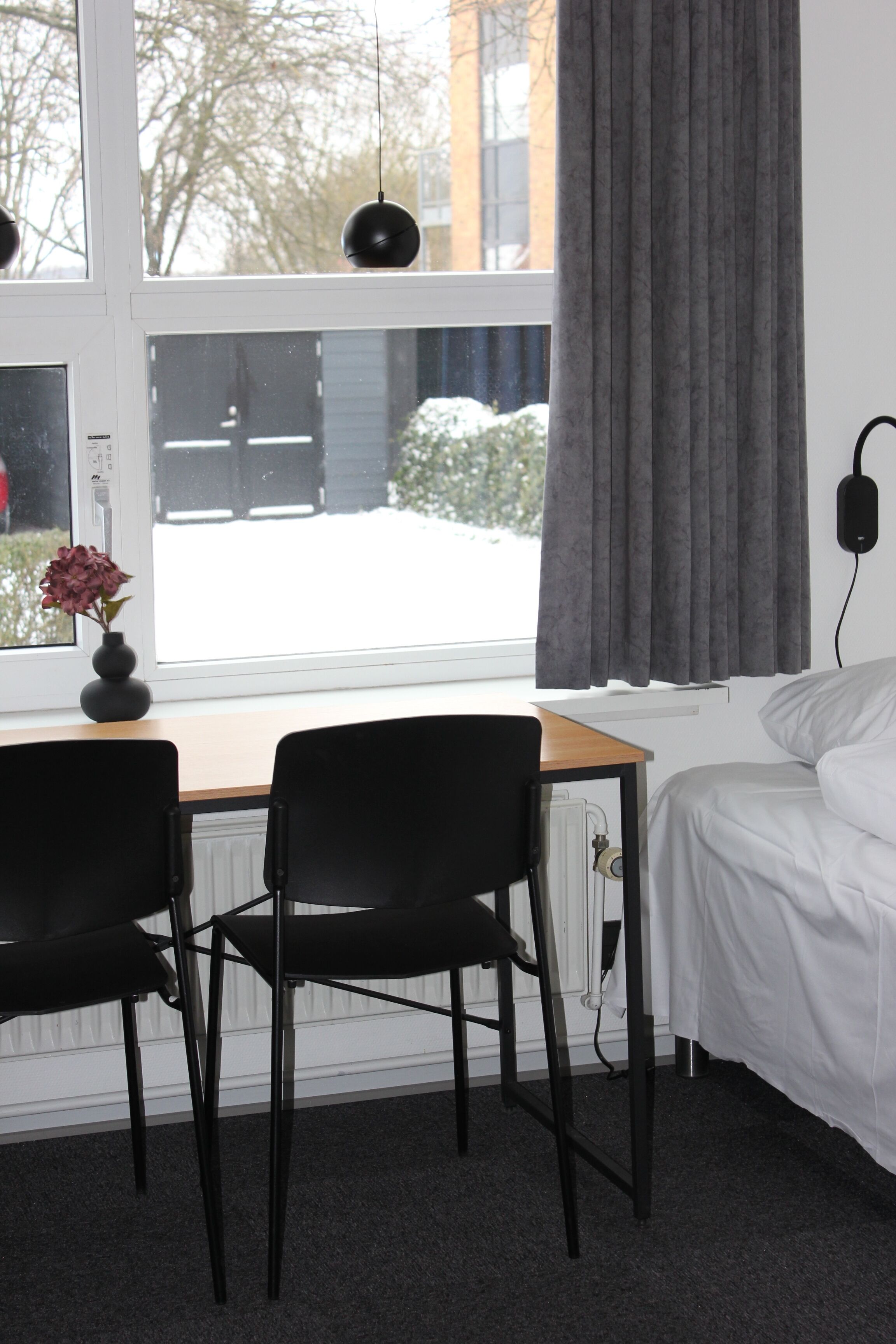 Un dormitorio con cama, un escritorio con dos sillas, una ventana con cortinas y un jarrón con flores.