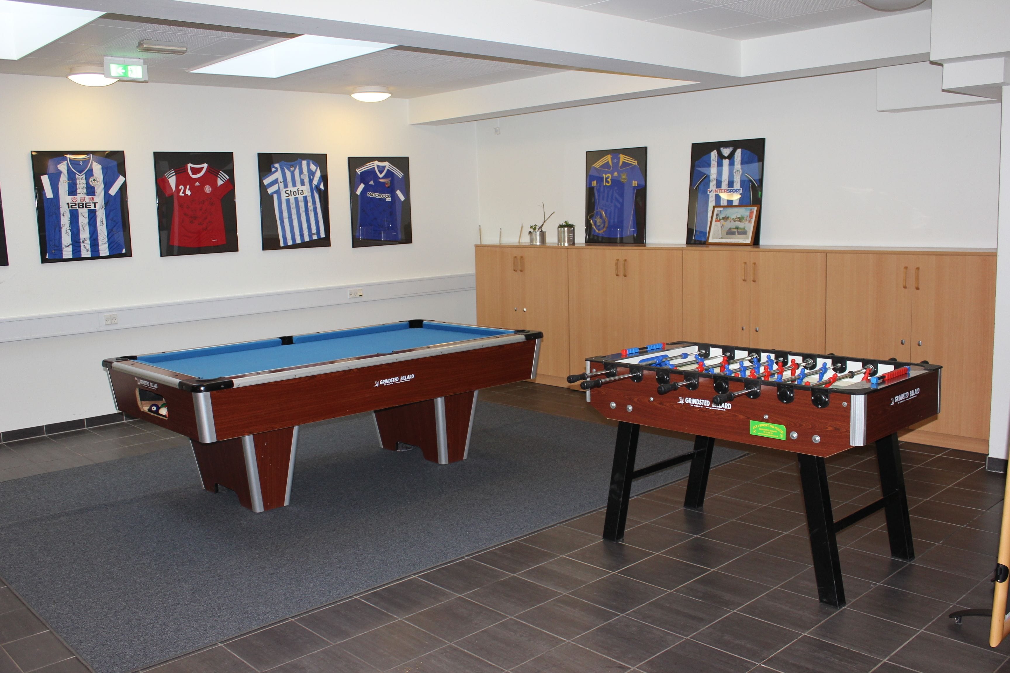 A pool table and a foosball table in a room with framed sports jerseys on the wall.