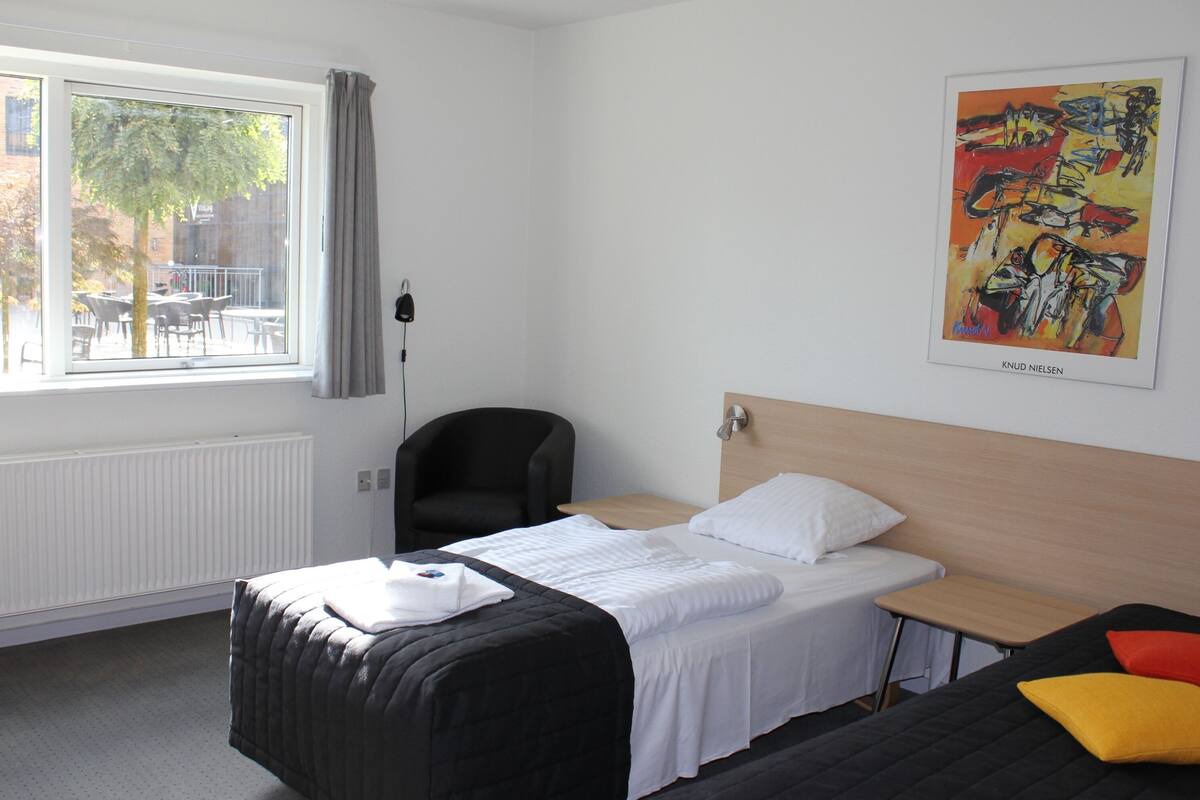 Una habitación de hotel con cama, escritorio, silla, ventana con vistas al exterior y un cuadro en la pared.