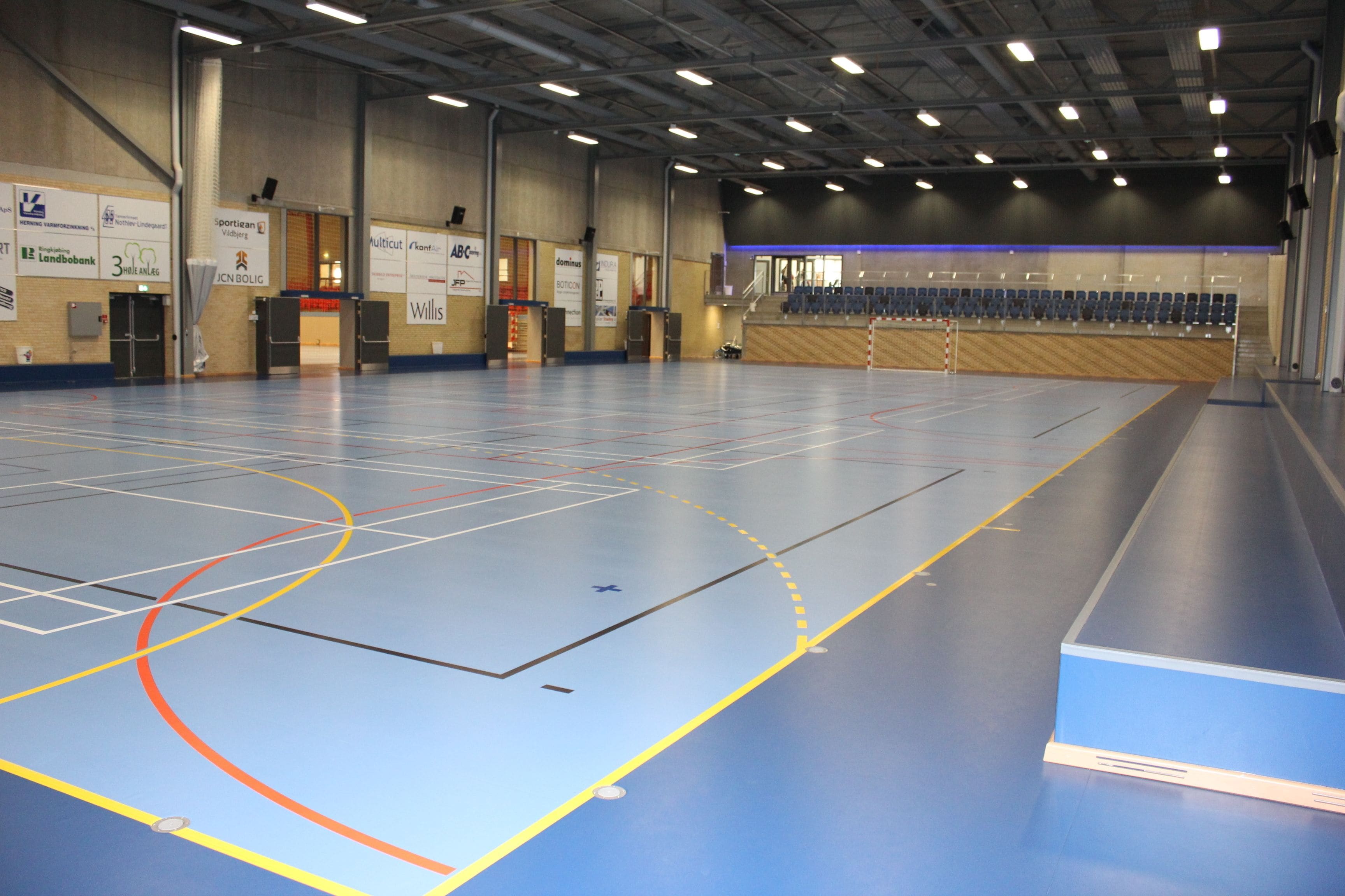 An indoor sports hall with a basketball court, seating area, and various banners on the walls.