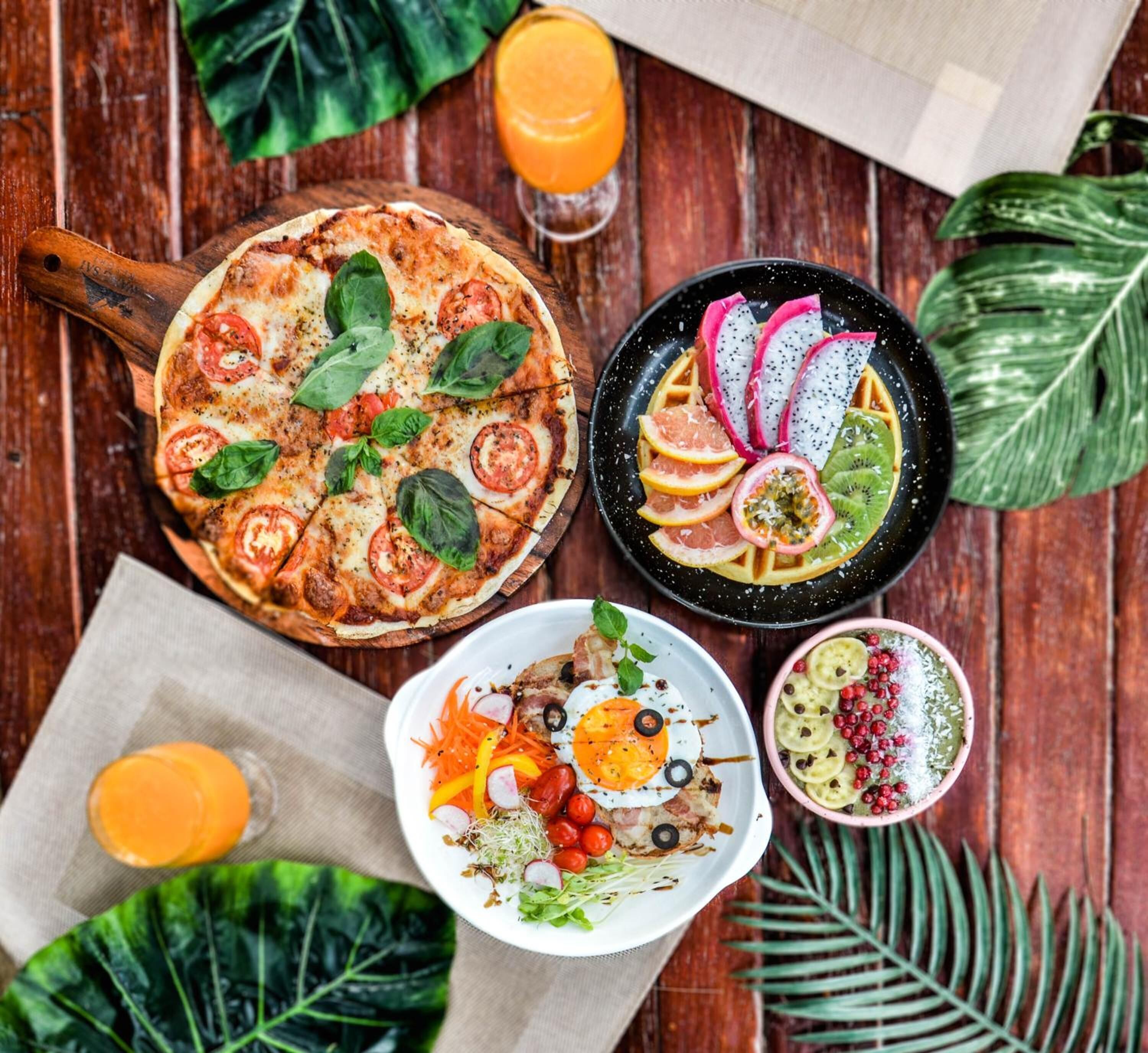 Une table en bois dressée pour le petit-déjeuner, avec une pizza, une salade de fruits et divers plats.