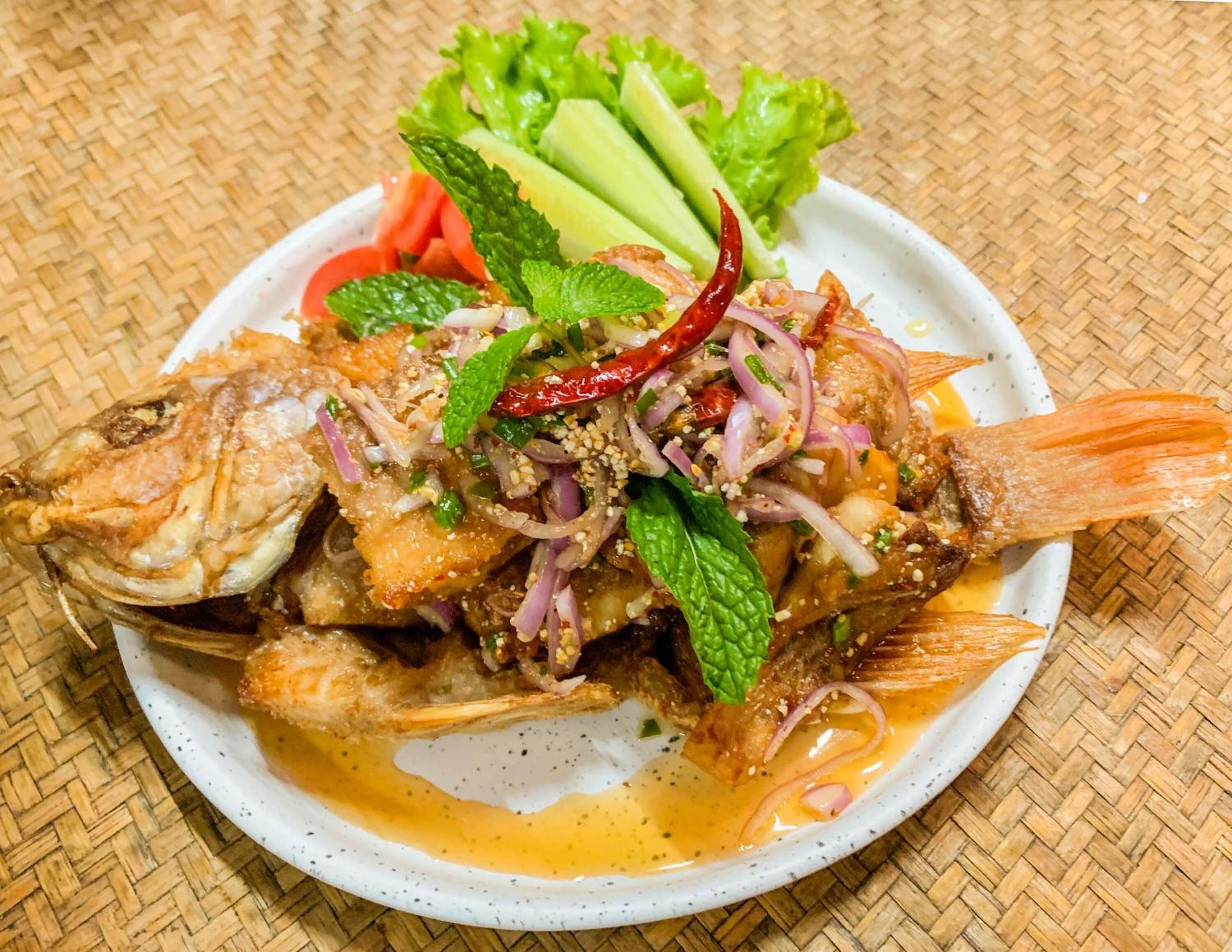 A plate of fried fish with a sauce, garnished with red chili, onions, and herbs, served on a woven mat.