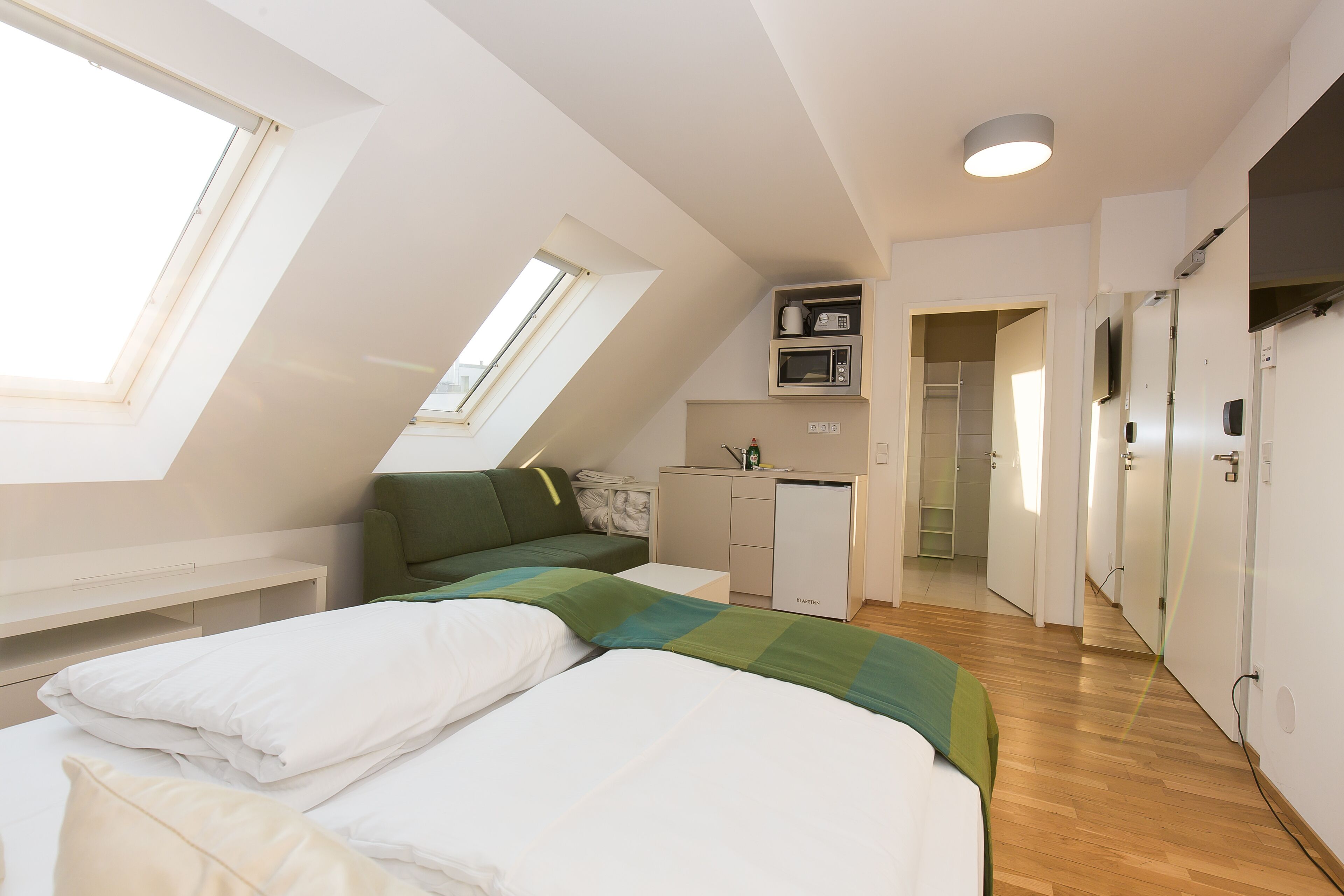 Een moderne loft slaapkamer met schuine plafonds, dakraam, een groene bank, een bed met witte lakens, een kitchenette en een inloopkast.