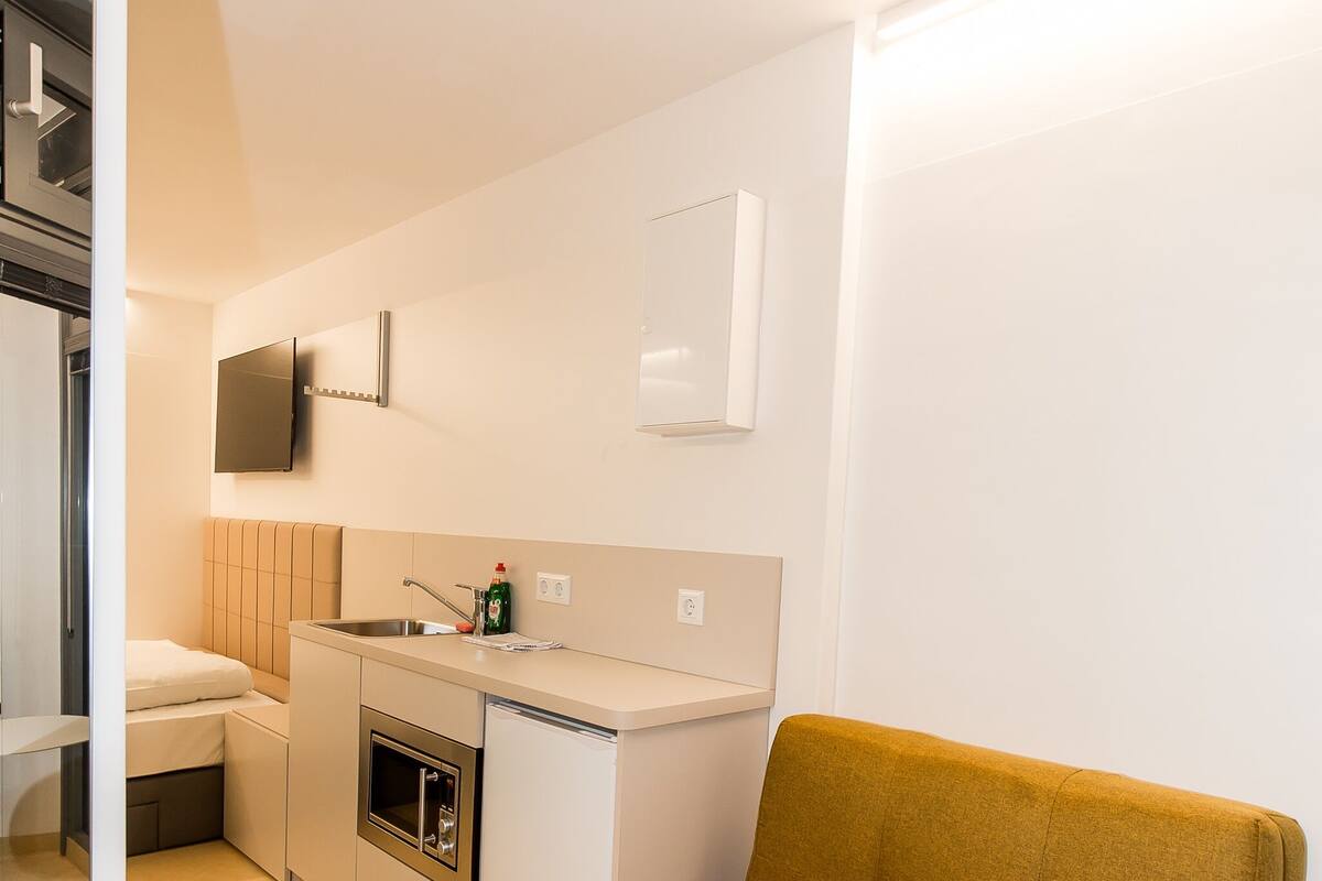 A modern kitchen with white cabinetry, a built-in oven, and a sink. There is a mustard-colored sofa in the foreground.