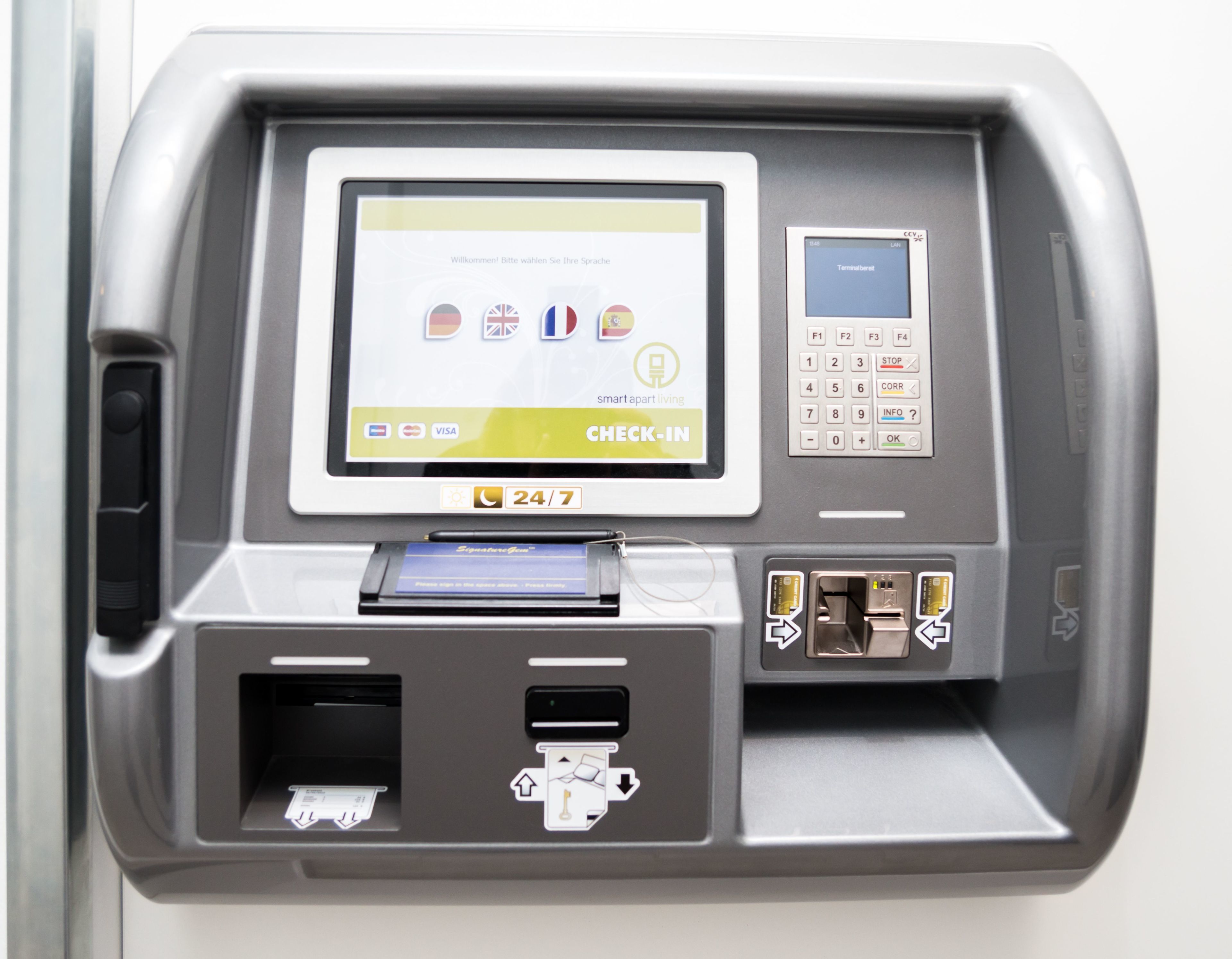 Un kiosque d’enregistrement en libre-service doté d’un écran affichant les options de langue, les logos Visa et Mastercard, et un indicateur de service disponible 24 heures sur 24, 7 jours sur 7.