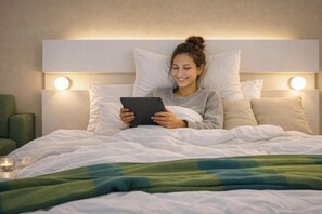 Una persona acostada en la cama con una tableta, una lámpara de noche y una silla verde.