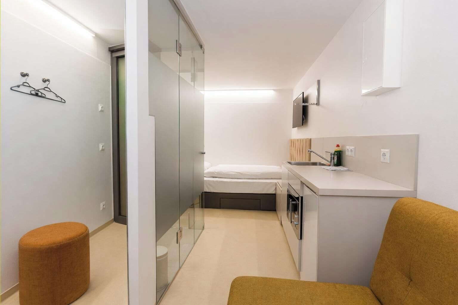 A compact hotel room with a bed, a kitchenette, a small stool, and a glass partition.