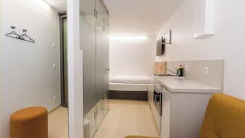 A compact hotel room with a bed, a kitchenette, a small stool, and a glass partition.