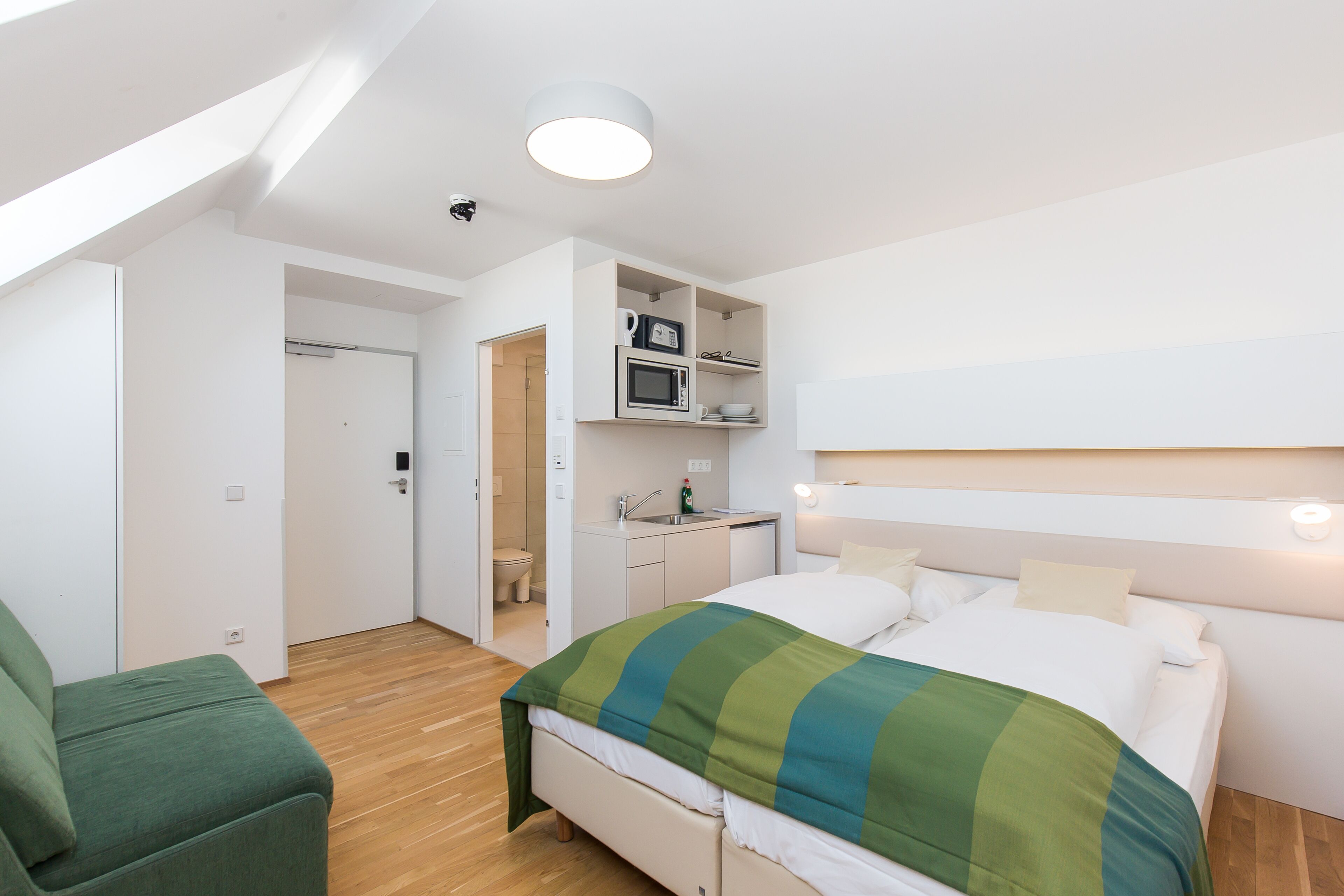 A modern hotel room with a bed, a green armchair, a kitchenette, and a bathroom.
