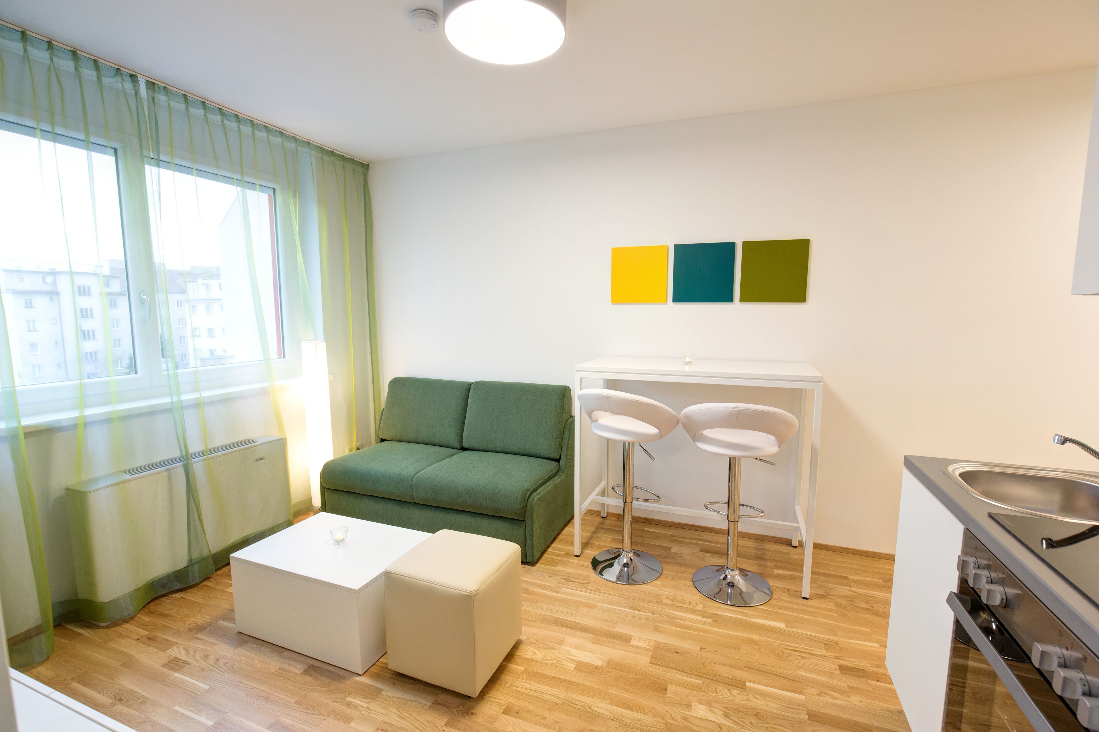 Una sala de estar moderna con un sofá verde, una mesa de centro blanca y dos taburetes de bar blancos. Incluye una zona de cocina con un fregadero y una estufa.