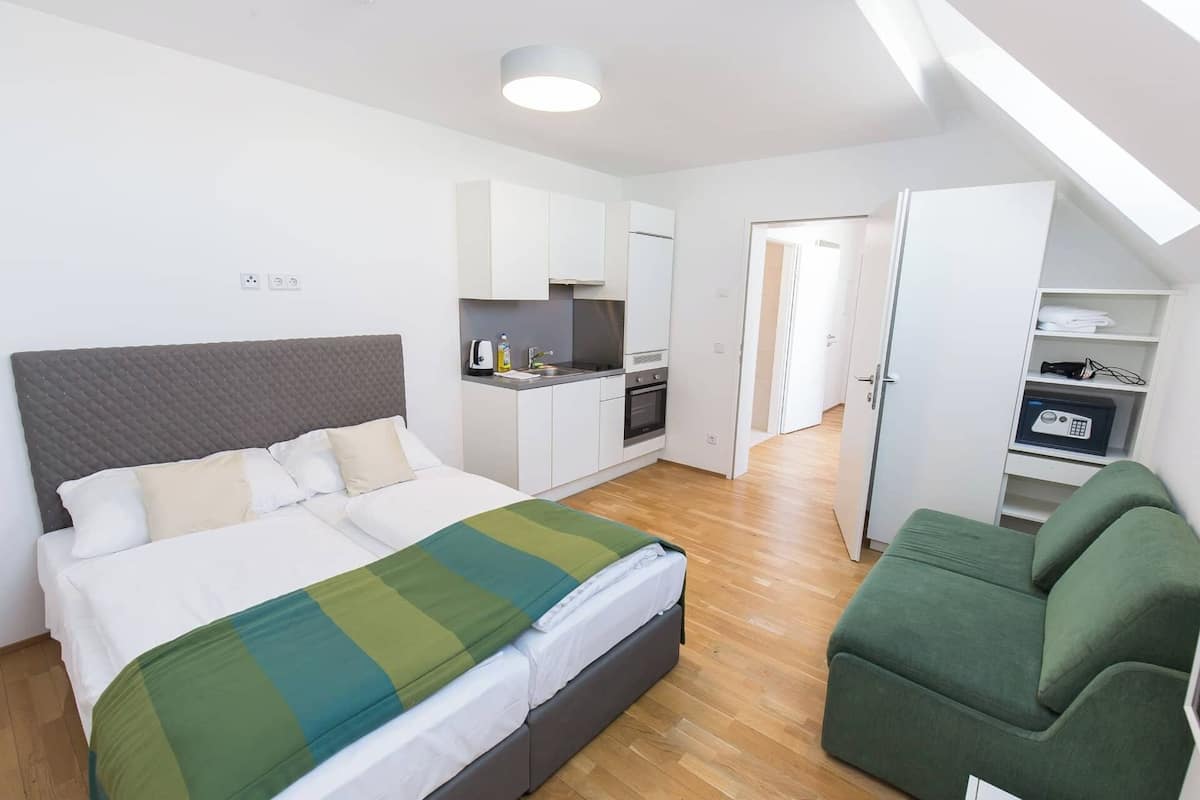 A modern hotel room with a bed, a green sofa, a kitchenette, and a walk-in closet.