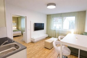 Ein modernes, gut beleuchtetes Zimmer mit einem Flachbildfernseher an einer weißen Wand, einer kleinen Küchenzeile mit Spüle, einem Essbereich mit weißem Tisch und Stühlen sowie einem Bett mit grüner Decke im Hintergrund.