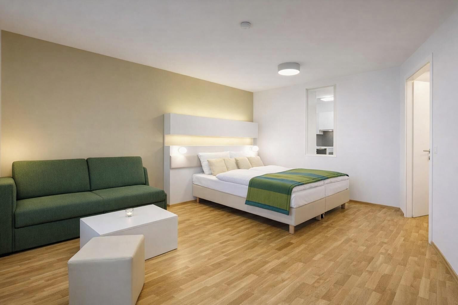A modern hotel room with a green sofa, a white bed with a green blanket, a white bedside table, and a wooden floor.