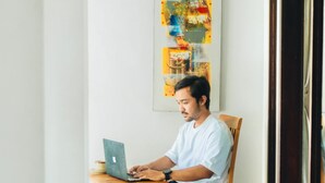 Een persoon zit aan een houten tafel met een laptop, in een ruimte met een witte muur en een kleurrijk abstract schilderij.