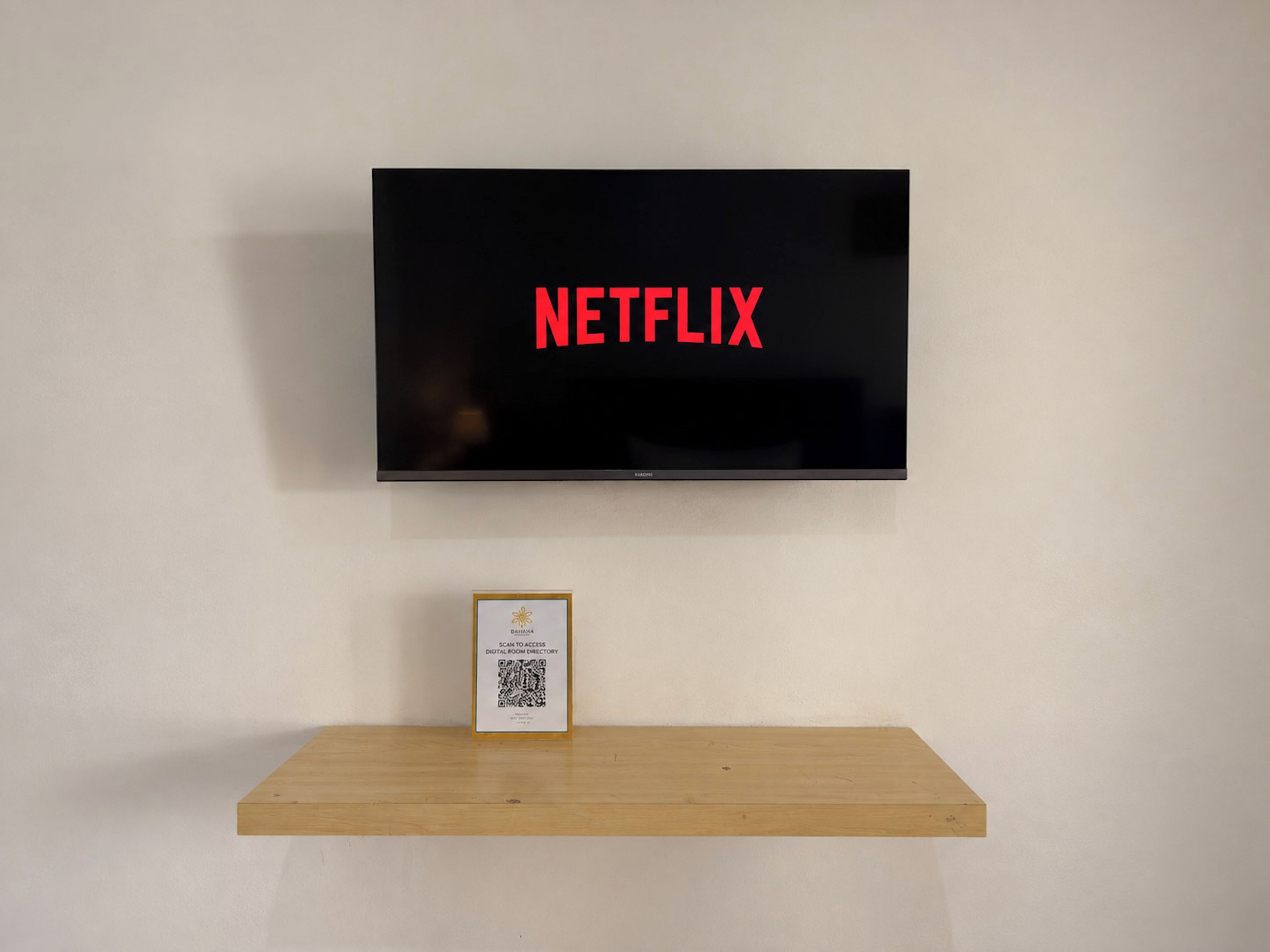 Seinälle asennettu litteänäyttöinen televisio, jossa sana »NETFLIX» on kirjoitettu punaisilla kirjaimilla. Television alapuolella on puinen hylly, jonka päällä on kehystetty esine ja QR-koodi.