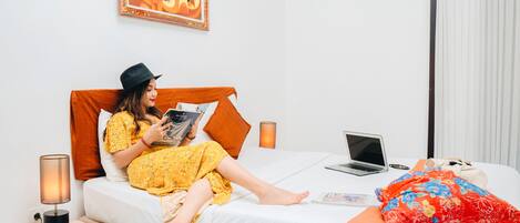 A person reading a book on a bed with a laptop, a lamp, and a pillow.
