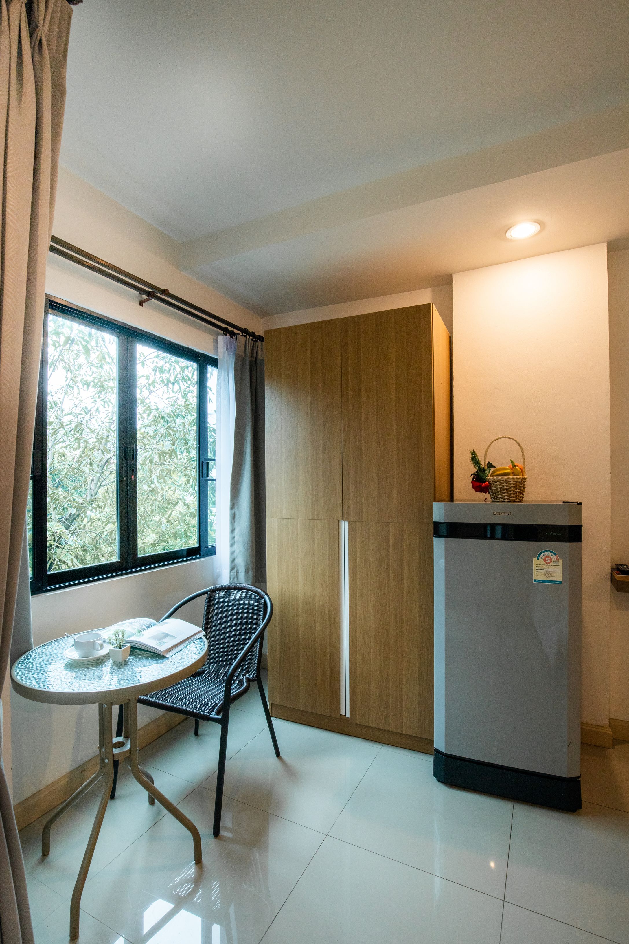 A compact room with a window, a table with a chair, a refrigerator, and a wooden wardrobe.