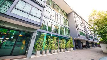 Un edificio moderno con fachada de vidrio, una zona de estacionamiento techada y una fila de macetas con plantas al frente.