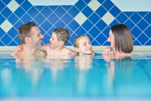 A family of four in a swimming pool with a blue and white tiled wall in the background. - Hotel Bellevue (Seeboden)