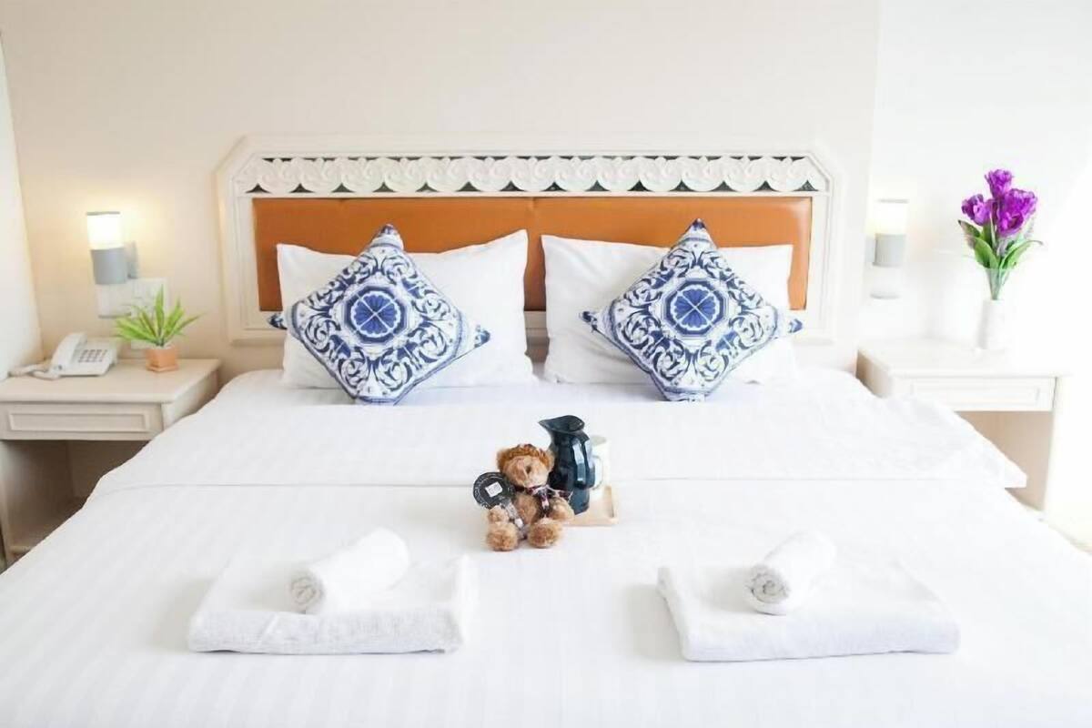 A neatly made bed with two decorative pillows, a small teddy bear, and a coffee pot on a bedside table.
