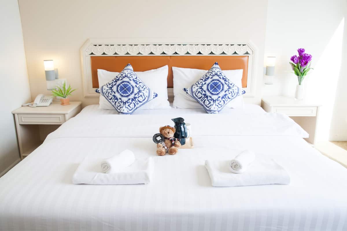 A neatly made bed with white linens, two decorative pillows, a teddy bear, a coffee pot, and two folded towels.