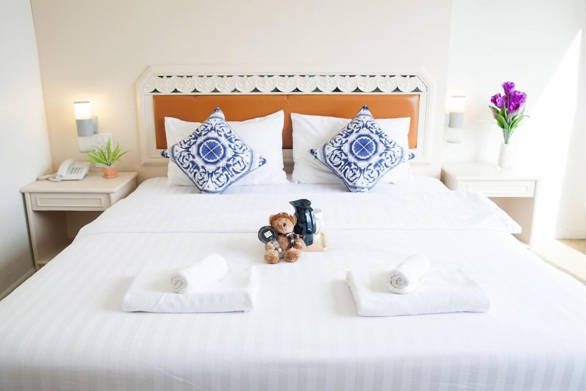 Quarto de hotel com cama bem arrumada, duas almofadas, um urso de pelúcia, uma cafeteira e um vaso com flores.