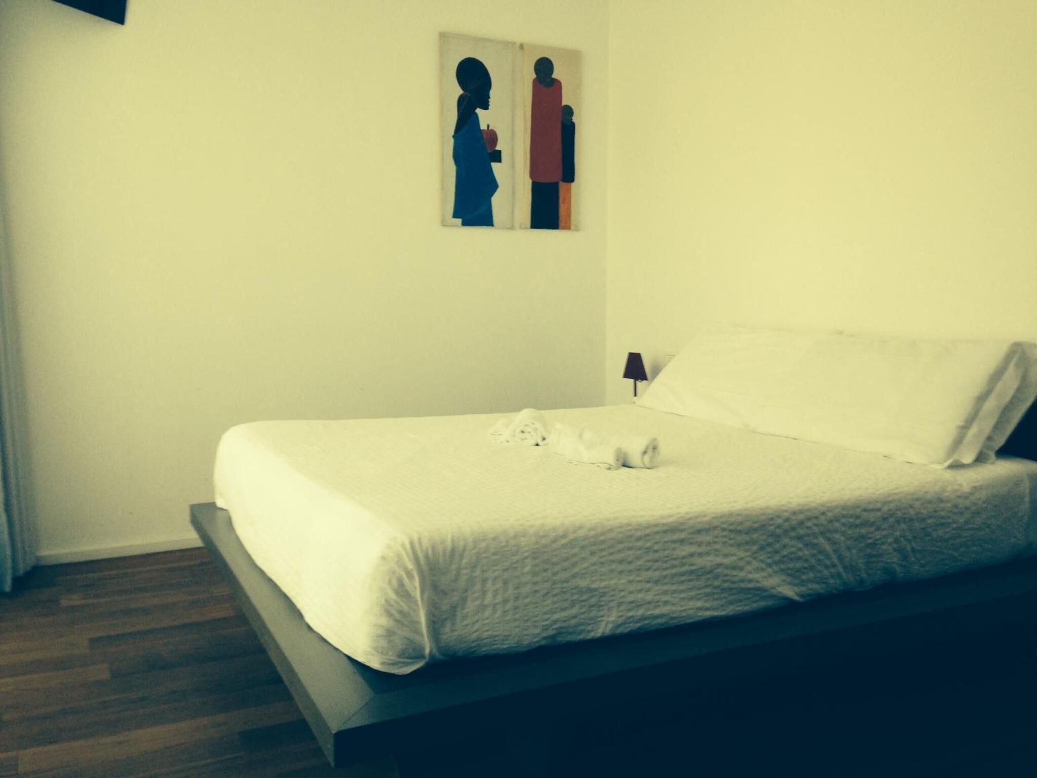Uma cama bem arrumada com edredom branco e almofadas, um abajur e uma obra de arte emoldurada na parede.