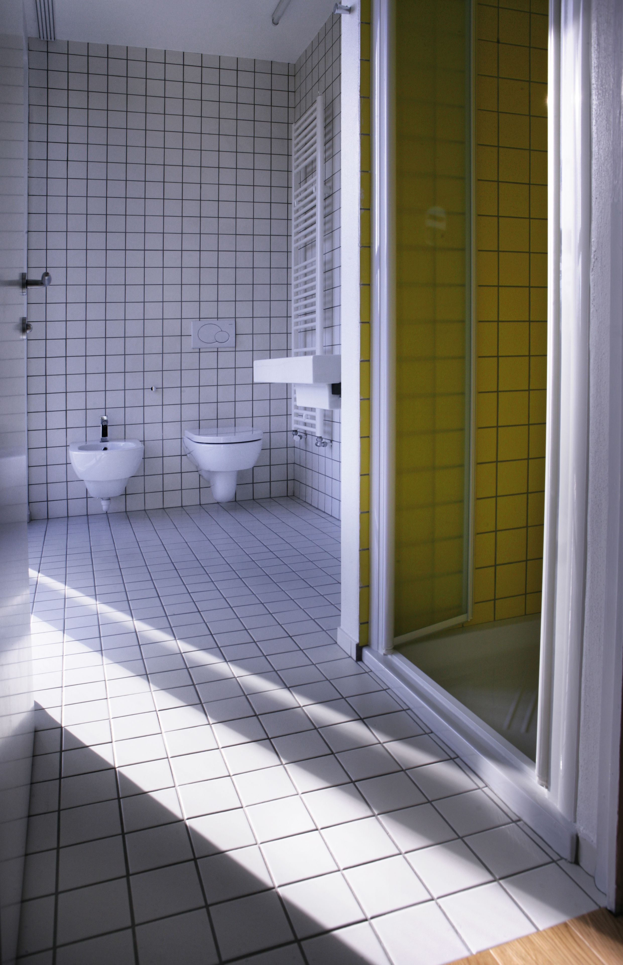 A bathroom with white tiled walls and floor, a white sink, a white toilet, and a yellow shower enclosure.