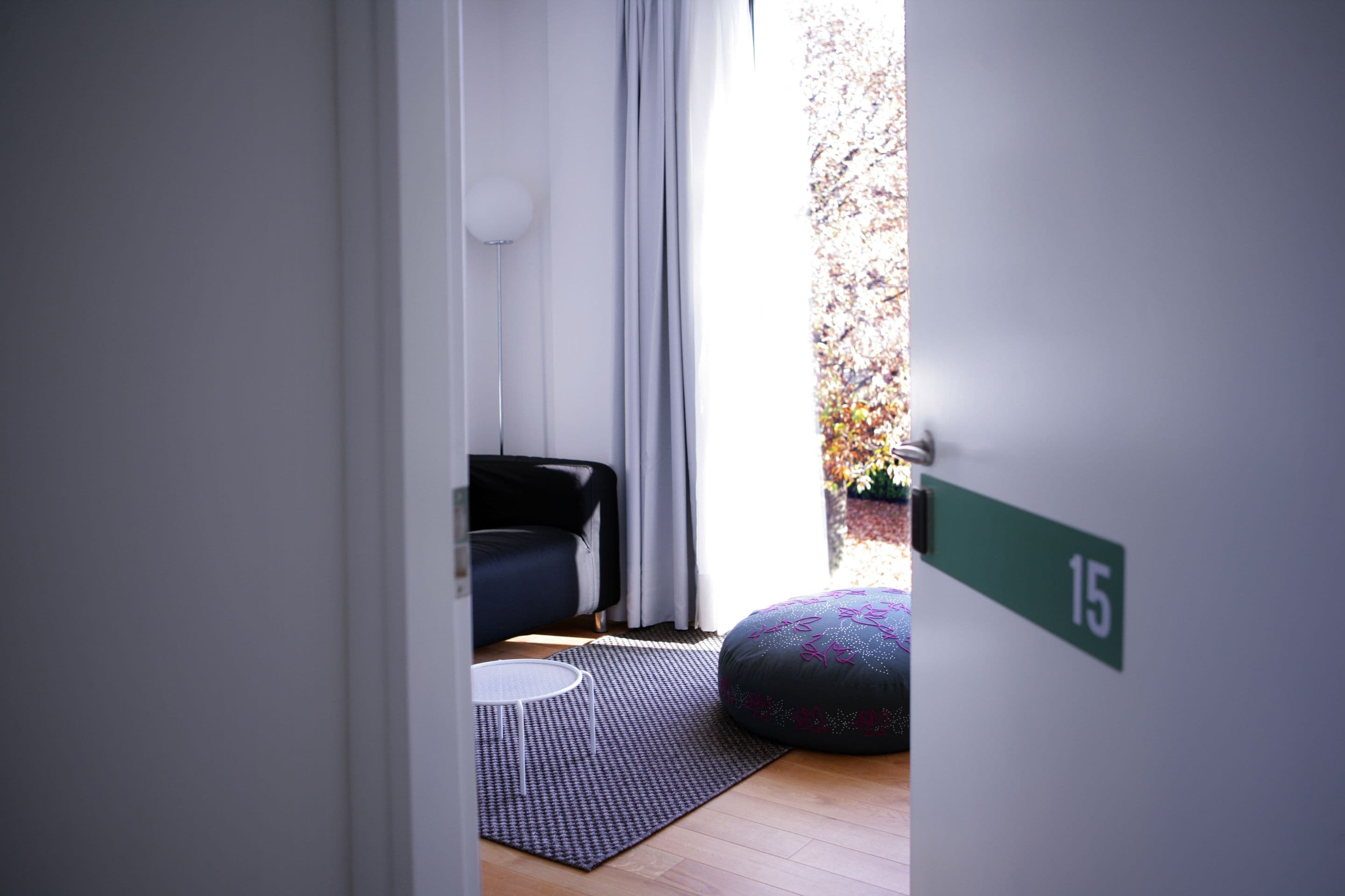 A modern room with a black sofa, a patterned ottoman, a small round table, and a view of a garden through a window.