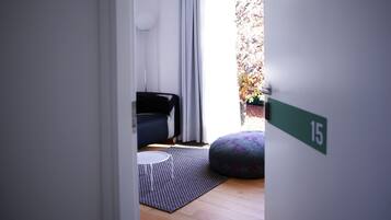 A modern room with a black sofa, a patterned ottoman, a small round table, and a view of a garden through a window.