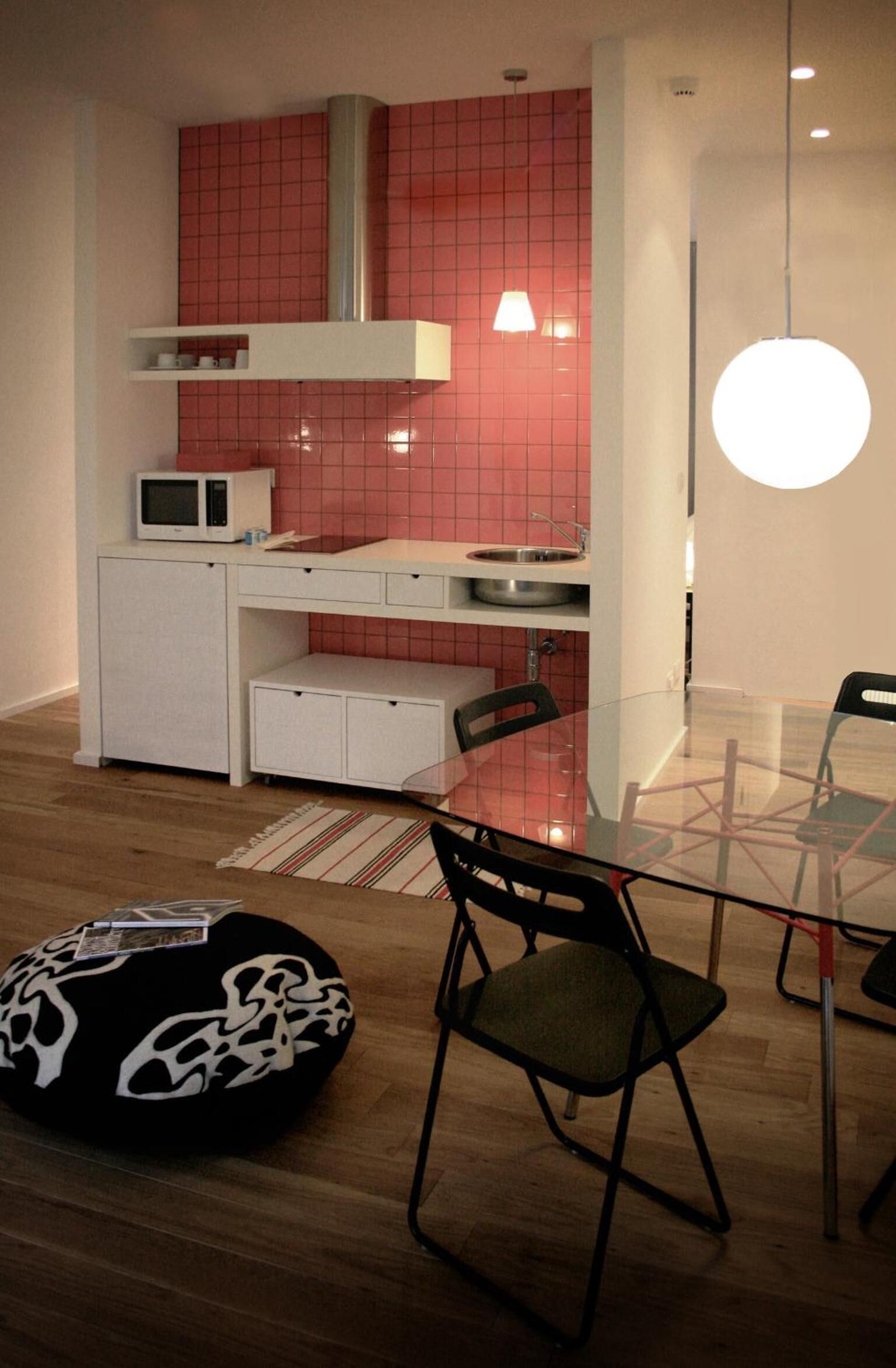Una cocina compacta con microondas, un lavamanos pequeño y un salpicadero de azulejos rojos. En primer plano, hay una mesa de comedor con sillas y una mesa con superficie de vidrio.