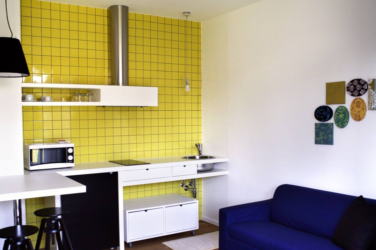 Een knus woongedeelte met een kitchenette, een blauwe bank en een gestreept voetenbankje.