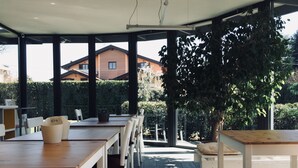 Un área de comedor al aire libre con mesas y sillas, un árbol grande y vistas a casas al fondo.